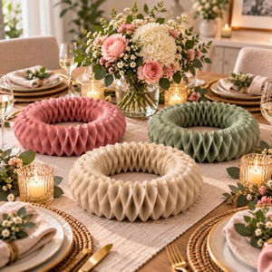 May include: Three circular honeycomb paper decorations in dusty rose, sage green, and cream colours arranged on a dining table. Each piece features a geometric accordion-folded structure, positioned among floral centrepieces and glass candle holders.