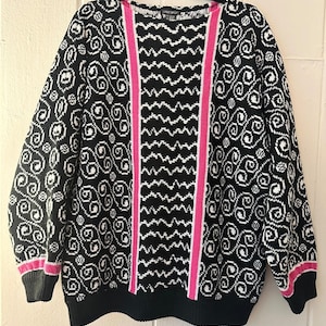 May include: A long-sleeve knit sweater featuring a black and white pattern of swirling circular motifs on the sides and a central vertical panel with a zigzag design. The garment includes bright pink vertical stripes and pink cuffs.