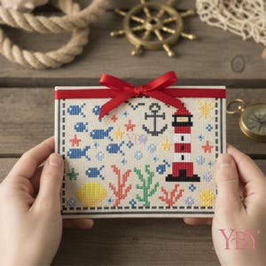 Puede incluir: Una pieza de bordado en punto de cruz rectangular sobre tela clara con temática náutica que incluye un faro rojo y blanco, un ancla negra, peces azules, coral y conchas marinas. Una cinta roja brillante está atada en un lazo arriba.