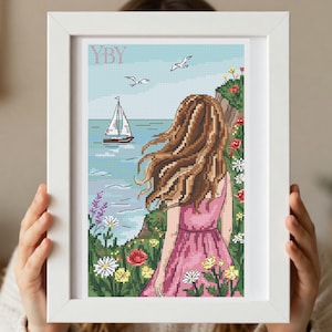 Puede incluir: Un bordado en punto de cruz enmarcado que muestra a una persona con cabello largo castaño y vestido rosa, de pie en un acantilado frente a un mar azul con un velero y dos aves. El texto YBY aparece impreso en la esquina superior izquierda.