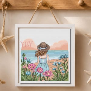 Puede incluir: Un patrón de punto de cruz enmarcado cuadrado que muestra a una persona con un vestido azul y un sombrero de sol marrón, de pie en una playa con flores rosas y azules en primer plano. El fondo muestra un mar tranquilo y un cielo al atardecer con las letras YBY.