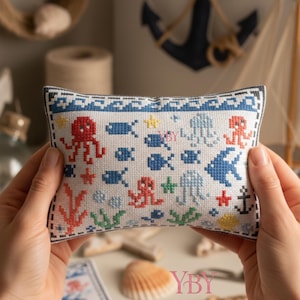 Puede incluir: Un acerico rectangular de punto de cruz con fondo blanco y borde de olas azules. El diseño incluye pulpos rojos, peces azules, estrellas de mar amarillas, algas verdes, medusas azul claro y un ancla negra, todo en un estilo pixelado.