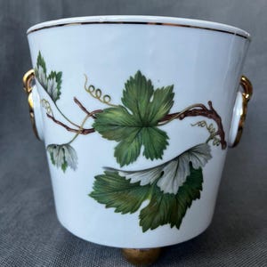 Puede incluir: Un cubo de hielo de porcelana blanca con un motivo pintado a mano de hojas de vid verdes con tallos marrones y asas de anillo doradas a cada lado. El recipiente tiene un borde dorado y una base de pedestal dorada en el centro.