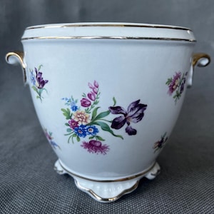 Op de afbeelding: Een witte porseleinen kom met een handgeschilderd bloemenpatroon in paars, blauw, geel en roze. De kom heeft twee goudkleurige handvatten aan de zijkanten, een goudkleurige rand en een geschulpte voet met gouden accenten.