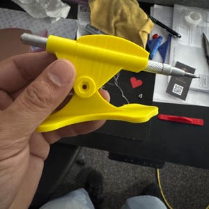 May include: A bright yellow 3D-printed stand featuring a curved base and a central pivot point, designed to hold a silver metal mechanical pencil or drafting tool securely in a horizontal position for display or easy access on a desk surface.