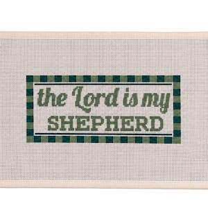 May include: A rectangular needlepoint canvas featuring a white mesh background with a central green and dark green checkered border. The text "the Lord is my SHEPHERD" is stitched in the center in a dark green, serif-style font.