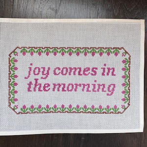 May include: A rectangular needlepoint canvas featuring the text "joy comes in the morning" stitched in pink thread. The text is enclosed within a decorative border composed of small green leaves and pink flower buds on a brown vine pattern.