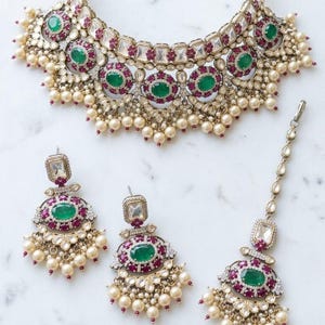 May include: A jewelry set consisting of a wide necklace, two drop earrings, and a maang tikka. Each piece features oval green stones surrounded by small red stones, clear crystals, and dangling cream-colored pearls set in a gold-toned metal frame.