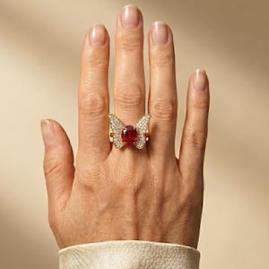 May include: A gold ring featuring a butterfly design with a central oval-cut red gemstone and small clear pavé-set stones covering the wings. The ring is worn on a finger, showcasing the detailed metalwork and the vibrant red centre stone.