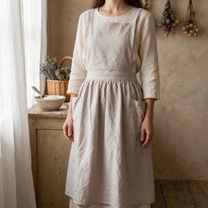 Op de afbeelding: Een lichtbeige linnen schort met een vierkante halslijn, brede schouderbanden en een geplooide rok met twee zijzakken. Het schort wordt gedragen over een bijpassend linnen shirt met lange mouwen voor een gecoördineerde, neutrale look.