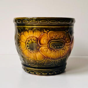 May include: A ceramic planter featuring a deep olive green glaze with prominent embossed golden-yellow floral motifs. The cylindrical vessel has a textured surface with a repeating circular pattern along the top and bottom rims and a glossy finish.