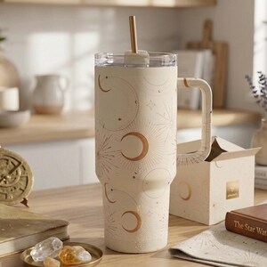 Puede incluir: Un vaso térmico de color crema con asa y pajita, que presenta un patrón celestial dorado de lunas y estrellas. El vaso tiene una tapa transparente y está decorado con un diseño repetitivo de lunas crecientes, soles y estrellas.