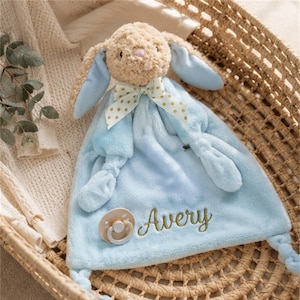 Peut inclure: Une couverture de sécurité pour bébé en peluche avec une tête de lapin beige aux longues oreilles doublées de bleu, un ruban blanc à pois dorés et un corps en tissu bleu clair doux. Le prénom Avery est brodé en fil doré et une sucette beige est fixée.