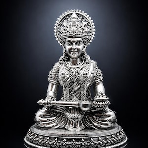Peut inclure: Une figurine métallique argentée représentant une divinité assise portant une couronne ornée et une tenue traditionnelle. La figure tient une louche dans la main droite et un petit bol dans la main gauche, sur une base circulaire détaillée.