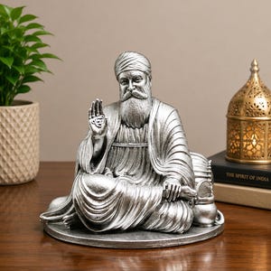 Könnte beinhalten: Eine silberfarbene Figur eines bärtigen Mannes mit Turban und traditionellem Gewand, der im Schneidersitz auf einem runden Sockel sitzt. Eine Hand ist mit offener Handfläche erhoben, die andere ruht auf einem kleinen Gefäß.