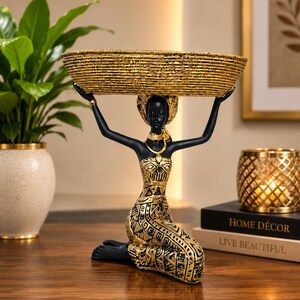 May include: A decorative figurine of a kneeling person with black skin and gold accents, wearing a patterned gold and black garment and headpiece, holding a large, textured, oval-shaped gold basket above their head with both raised arms.