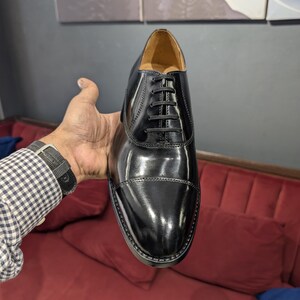May include: A polished black leather oxford shoe featuring a cap toe design, a closed lacing system with thin black laces, and a tan interior lining. The shoe has a sleek, smooth finish with visible stitching along the cap toe and eyelet panels.