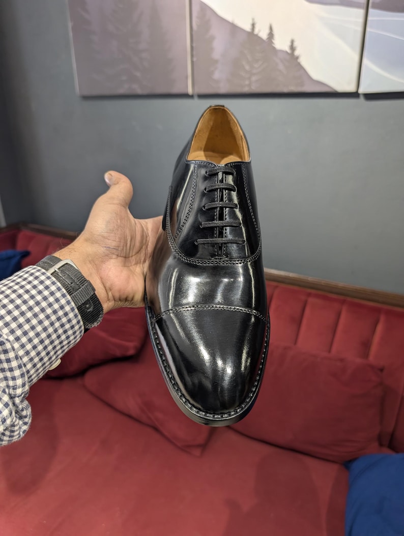 May include: A polished black leather oxford shoe featuring a cap toe design, a closed lacing system with thin black laces, and a tan interior lining. The shoe has a sleek, smooth finish with visible stitching along the cap toe and eyelet panels.