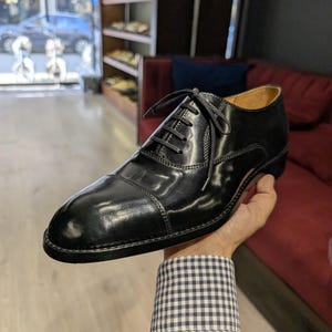 May include: A polished black leather oxford shoe featuring a classic cap toe design, a closed lacing system with thin black laces, and a low heel. The shoe has visible stitching along the cap toe and side panels, with a smooth, shiny finish.