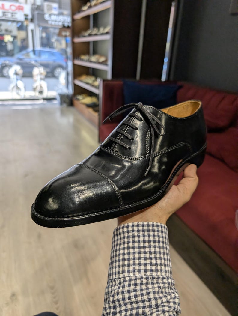 May include: A polished black leather oxford shoe featuring a classic cap toe design, a closed lacing system with thin black laces, and a low heel. The shoe has visible stitching along the cap toe and side panels, with a smooth, shiny finish.