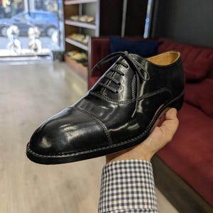 May include: A black leather oxford shoe with a cap toe design, featuring a polished finish, thin black laces, and a low heel. The shoe has visible stitching along the side panels and a tan interior lining, showcasing a classic formal silhouette.