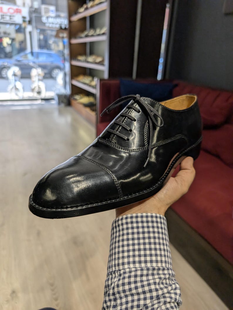 May include: A black leather oxford shoe with a cap toe design, featuring a polished finish, thin black laces, and a low heel. The shoe has visible stitching along the side panels and a tan interior lining, showcasing a classic formal silhouette.