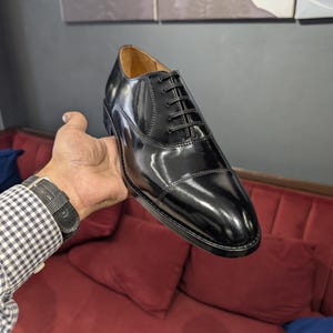 May include: A polished black leather oxford shoe featuring a cap toe design, a closed lacing system with thin black laces, and a tan leather interior lining. The shoe has a sleek, glossy finish and a sturdy black sole with visible stitching.