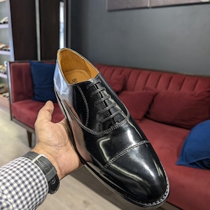 May include: A polished black leather oxford shoe featuring a cap toe design, thin black laces, and a tan interior lining. The shoe has a sleek, glossy finish with visible stitching along the cap toe and eyelet panels, showcasing a classic silhouette.