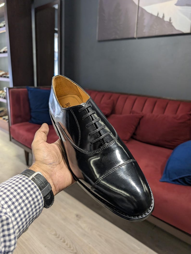 May include: A polished black leather oxford shoe featuring a cap toe design, thin black laces, and a tan interior lining. The shoe has a sleek, glossy finish with visible stitching along the cap toe and eyelet panels, showcasing a classic silhouette.