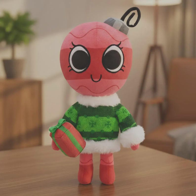 May include: A plush toy shaped like a round pink and red Christmas ornament with a grey cap and a black loop. The figure wears a green knit sweater with white faux fur trim at the collar and cuffs, and holds a small red gift with a green ribbon.
