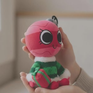 May include: A plush toy shaped like a round pink Christmas ornament with a smiling face, large black eyes, and eyelashes. It wears a green knitted sweater with white faux fur trim at the collar and cuffs, and holds a small red and green gift box.