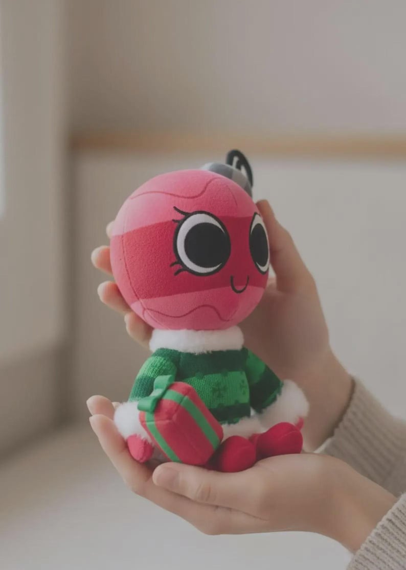 May include: A plush toy shaped like a round pink Christmas ornament with a smiling face, large black eyes, and eyelashes. It wears a green knitted sweater with white faux fur trim at the collar and cuffs, and holds a small red and green gift box.