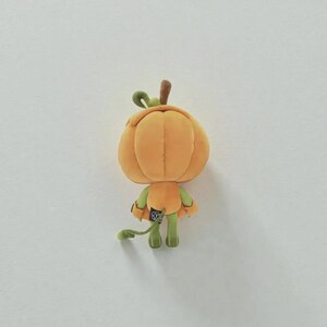 以下が含まれることがあります： 茶色の茎と緑色の手足を持つ、オレンジ色のカボチャの形をしたぬいぐるみ。カボチャはセグメント化された体、細い緑色の腕と脚、背面に小さな黒いタグが付いています。おもちゃは後ろから見た視点です。