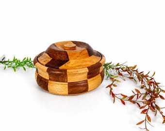 Puede incluir: Un recipiente redondo de madera con tapa, que presenta un patrón de tablero de ajedrez en contraste de madera clara y marrón oscuro. El recipiente tiene un acabado liso y pulido y un pequeño pomo circular en el centro de la tapa.