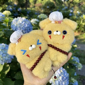 May include: Two yellow plush bear-like figures with fuzzy texture, each wearing a small white chef hat with a red and white checkered accent. They feature dark eyes, white bead noses, red triangular cheek markings, and a string of dark red beads.
