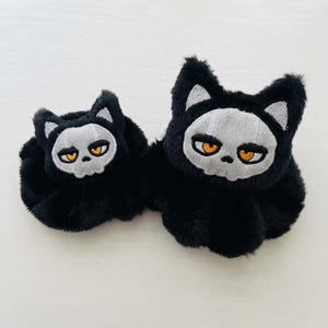 May include: Two black plush hair scrunchies featuring an embroidered patch of a cat face with a skull-like appearance. The cat design has white ears, a light gray skull face, and bright orange eyes. The scrunchies are made of soft, fuzzy material.
