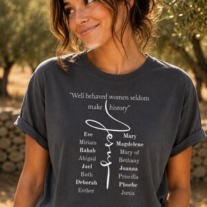 May include: A charcoal gray short-sleeve t-shirt featuring the white text "Well behaved women seldom make history" above a stylized "Jesus" script. Below the script are two columns of names: Eve, Miriam, Rahab, Abigail, Jael, Ruth, Deborah, Esther, Mary Magdalene, Mary of Bethany, Joanna, Priscilla, Phoebe, and Junia.