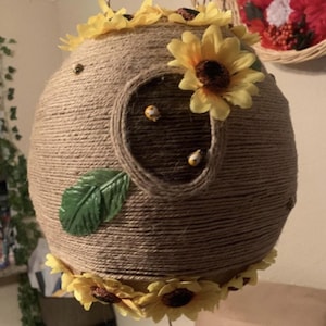 May include: A spherical hanging decoration wrapped in natural tan jute twine, featuring a circular opening. The surface is adorned with yellow artificial sunflower blossoms, two small green fabric leaves, and several tiny bee-shaped embellishments.