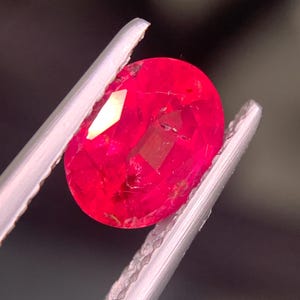 Może przedstawiać: Wibrujący rubin o szlifie owalnym, o głębokim czerwonym odcieniu z widocznymi inkluzjami wewnętrznymi, trzymany pewnie przez srebrzystą metalową pęsetę. Fasetowana powierzchnia kamienia odbija światło, podkreślając jego nasycony i półprzezroczysty kolor.