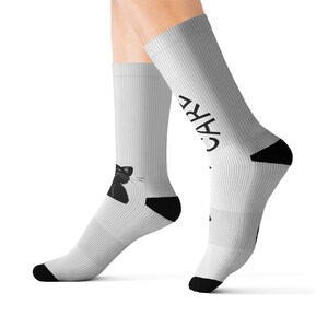 May include: A pair of white crew socks featuring a black graphic of a cat head on the lower side and the black text "CARE" printed vertically along the upper leg section. The socks have solid black heels and toe sections for contrast.