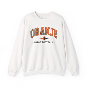 May include: A white long-sleeve crewneck sweatshirt featuring the text ORANJE in an arched orange font with a black outline. Below the text is a black and orange diamond-shaped icon and the words TOTAL FOOTBALL in a black sans-serif font.