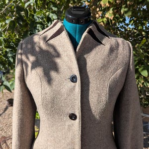 May include: A tan, textured wool-blend blazer featuring a high collar with dark brown velvet accents, long sleeves, and three black buttons down the front. The garment is displayed on a teal mannequin torso against a natural outdoor background.