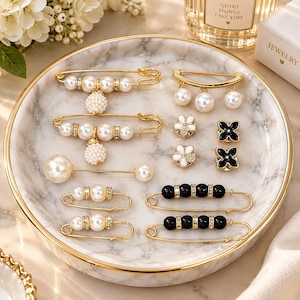 May include: A collection of gold-toned jewellery pins arranged on a round marble tray. The set includes safety pins adorned with white faux pearls, clear crystals, and black beads, alongside small flower-shaped and four-leaf clover-shaped pins.