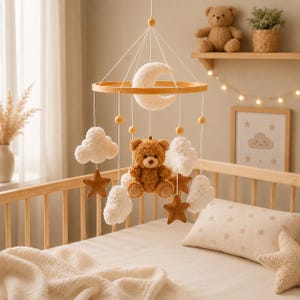 May include: A nursery mobile featuring a central brown teddy bear, a white crescent moon, white fluffy clouds, and brown stars suspended from a circular wooden frame by white strings with small wooden beads, designed for a crib setting.