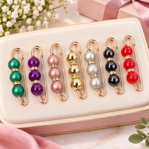 May include: A set of seven gold-toned safety pins, each featuring three round beads in green, purple, pink, gold, silver, black, and red. Each bead is separated by a small, clear rhinestone spacer, creating a decorative accessory set.