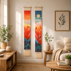 May include: Two vertical fabric wall hangings featuring mountain landscapes. The left piece displays a sunset gradient with warm oranges and reds. The right piece shows a sun against a blue sky. Both hang from wooden rods with cream tassels.