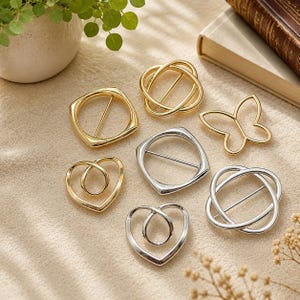 May include: A collection of seven metal belt buckles in gold and silver tones. The shapes include square, heart, butterfly, and circular interlocking designs. Each buckle features a central horizontal bar for fastening, with smooth, polished surfaces.
