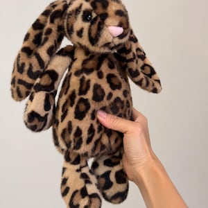 May include: A plush stuffed animal in the shape of a rabbit with long, floppy ears. The toy is covered in a soft, tan-colored faux fur fabric featuring a dark brown and black leopard print pattern. It has a small, light pink nose and dark eyes.