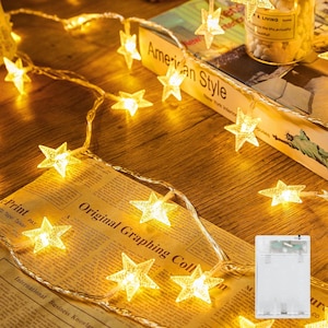 May include: A string of LED fairy lights featuring translucent, five-pointed star covers that emit a warm yellow glow. The lights are connected by a thin, clear wire and include a rectangular, transparent battery pack for power operation.