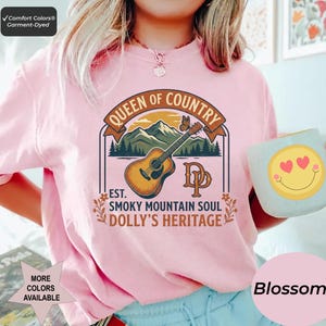 K&ouml;nnte beinhalten: Ein hellrosa T-Shirt mit einem Grafikdruck einer akustischen Gitarre, Bergen und einem Schmetterling. Der Text lautet QUEEN OF COUNTRY, EST. SMOKY MOUNTAIN SOUL, DOLLY'S HERITAGE und DP. Ein hellblauer Becher mit einem gelben Smiley wird gehalten.